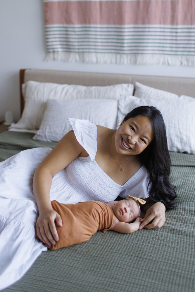 Newborn Photography, mother is lying on the bed next to her sleeping newborn baby girl smiling at the camera