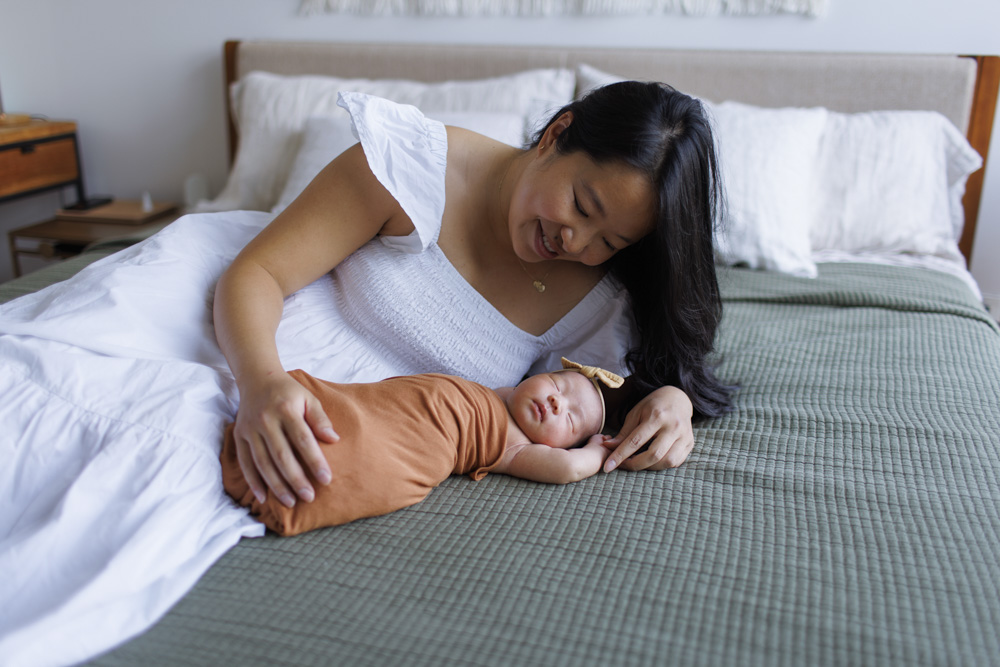Newborn Session, mother is lying on the bed next to her newborn baby girl, newborn wrapped up in a blanket wearing a headband is lying on its back with arm arms above her head while mom is looking at her