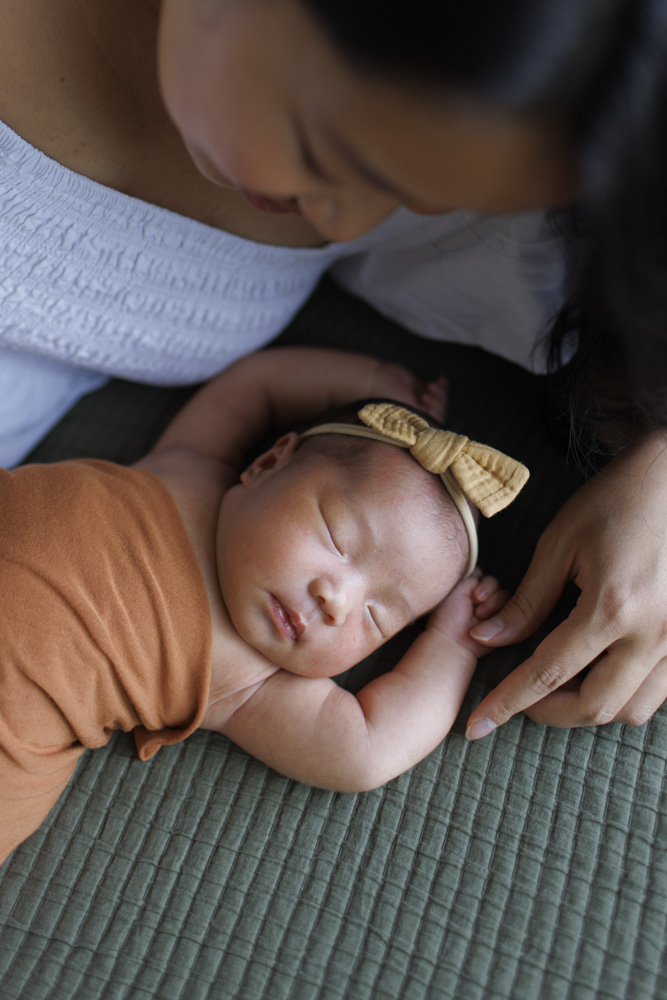 Baby Photography, close up of newborn wrapped up in a blanket wearing a headband sleeping on its back with arms above the head lying on a green blanket while mother is looking at her