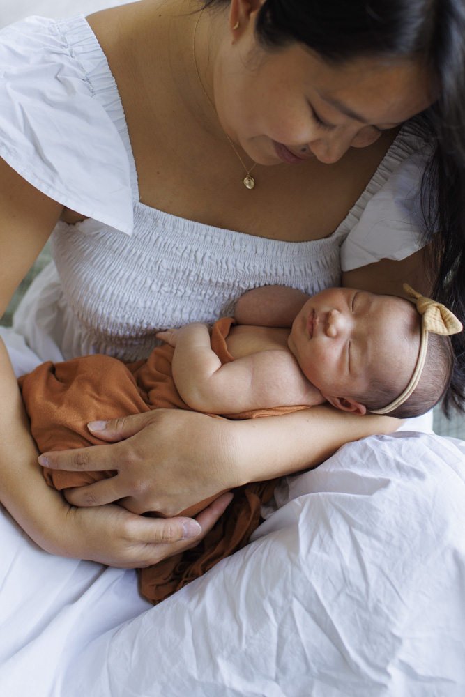 Newborn Photography, close up of mother holding her sleeping newborn baby in her arms, baby is wearing a cute headband with a bow and is wrapped up in a blanket