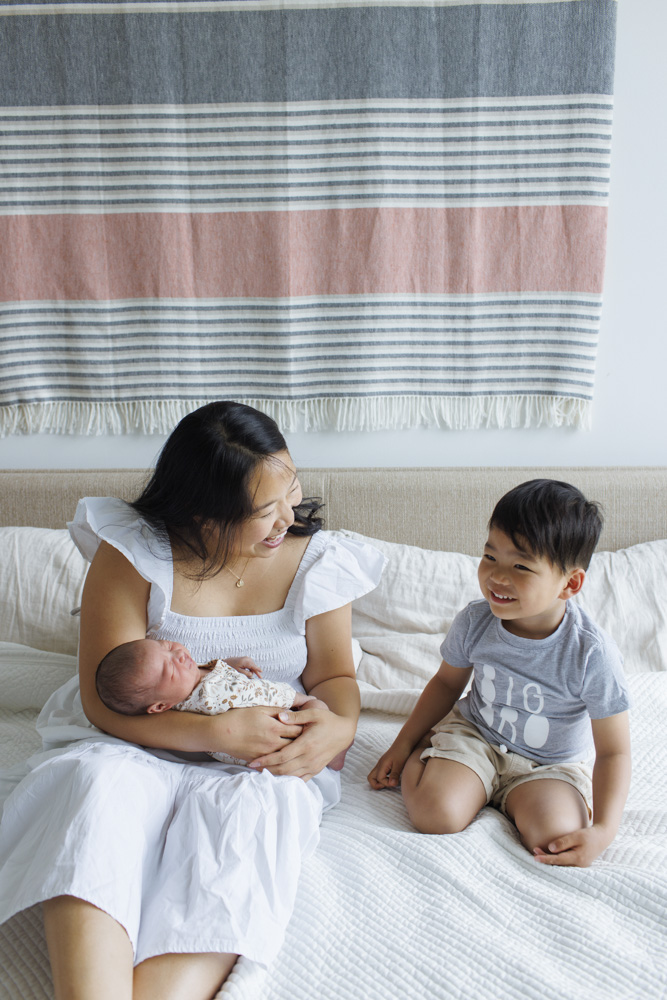 Family Session, mother sitting on a bed holding her sleeping newborn smiling at her toddler who is sitting next to her