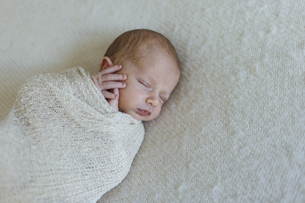 Baby Photography, close up of newborn lying on a beige sheet swaddled in a beige blanket with its hand sticking out eyes closed