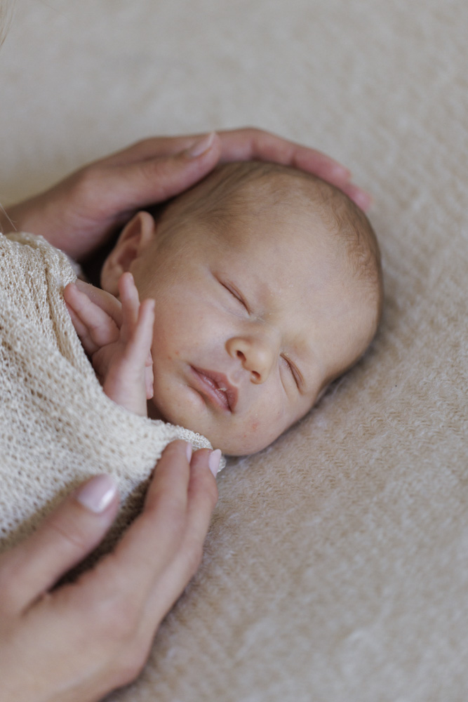 Baby Photography, close up of a sleeping newborn baby lying on a beige sheet swaddled in a beige blanket while mothers hands are touching it