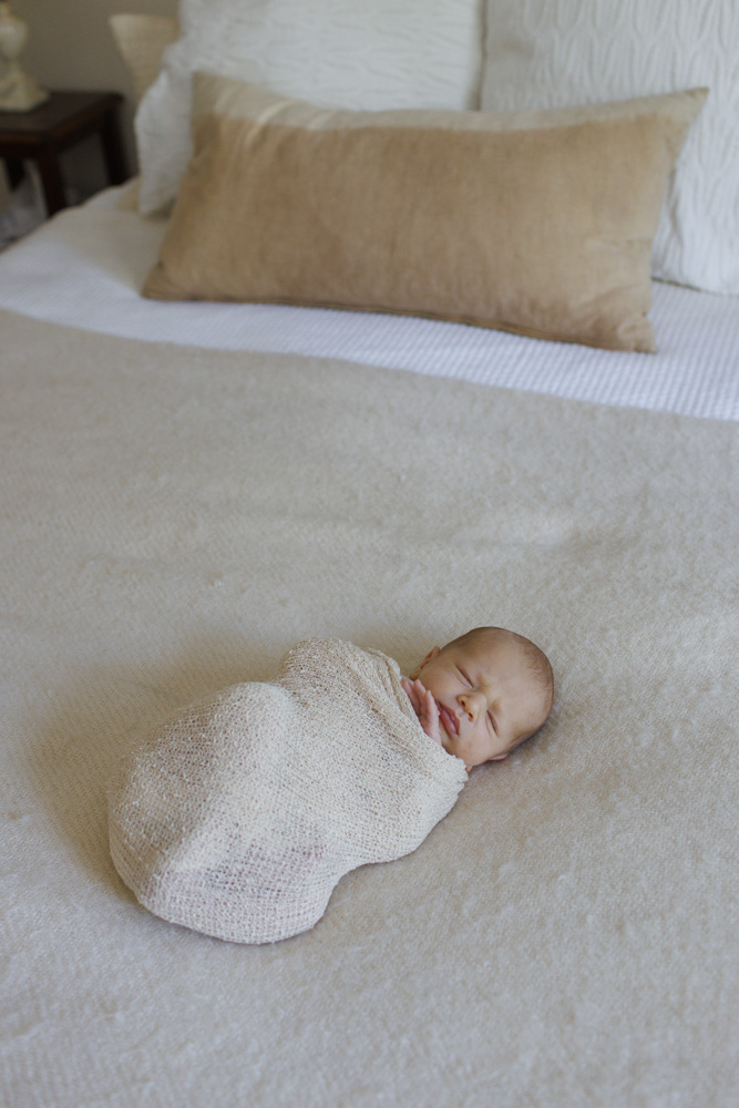 Baby Photography, newborn lying on a beige sheet swaddled in a beige blanket with its hand sticking out eyes closed