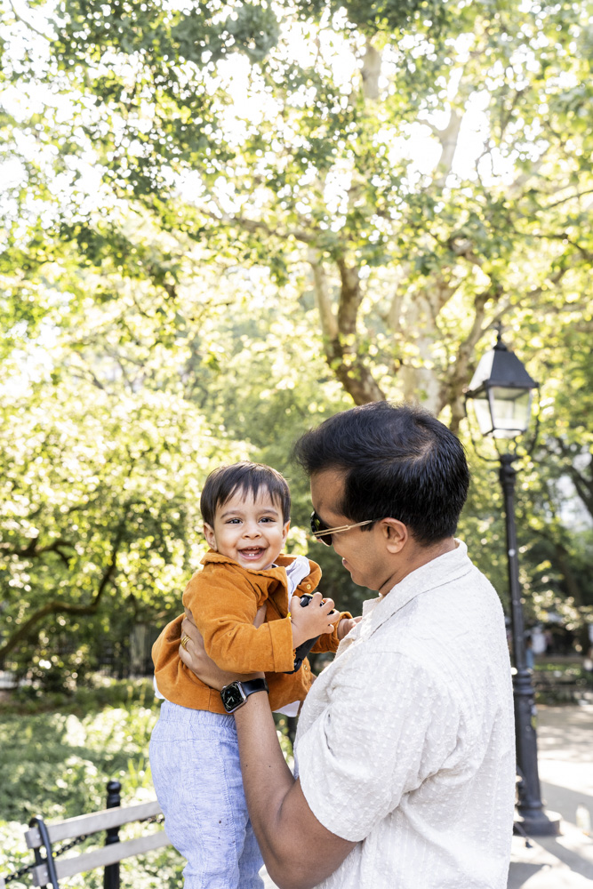 Family Session, father is holding his smiling toddler up, the background is a beautiful park