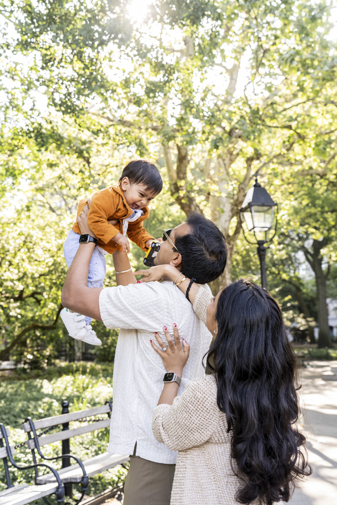 Family Session, father is holding his toddler up in the air while mom is standing behind dad smiling at them, the background is a beautiful park