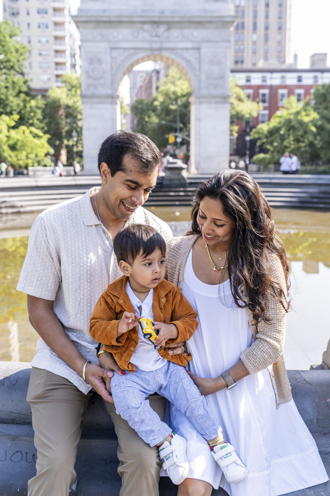 Family Photos, family of three sitting on the edge of a fountain, toddler is sitting between his parents holding a yellow toy car while parents are smiling at him