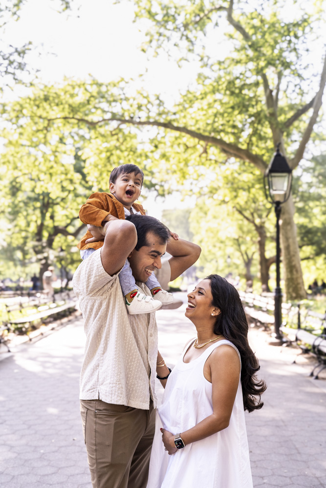 Family Photos, family of three is standing in a park. toddler is sitting on father's shoulder, mother is holding her belly and is looking at them smiling