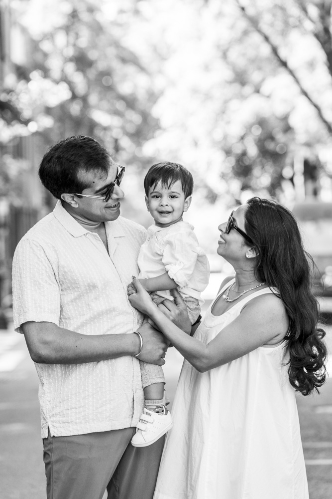 Family Photos, black and white photo of a family of three standing outside, father is holding his toddler while mother is looking at her son holding his hand, all are smiling