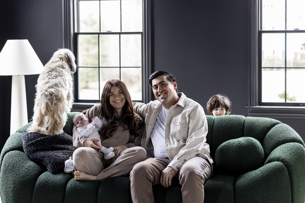 Family Session, parents are sitting on a green couch mom is holding the baby while dog is standing next to them looking the other direction and their toddler is peeking over the couch