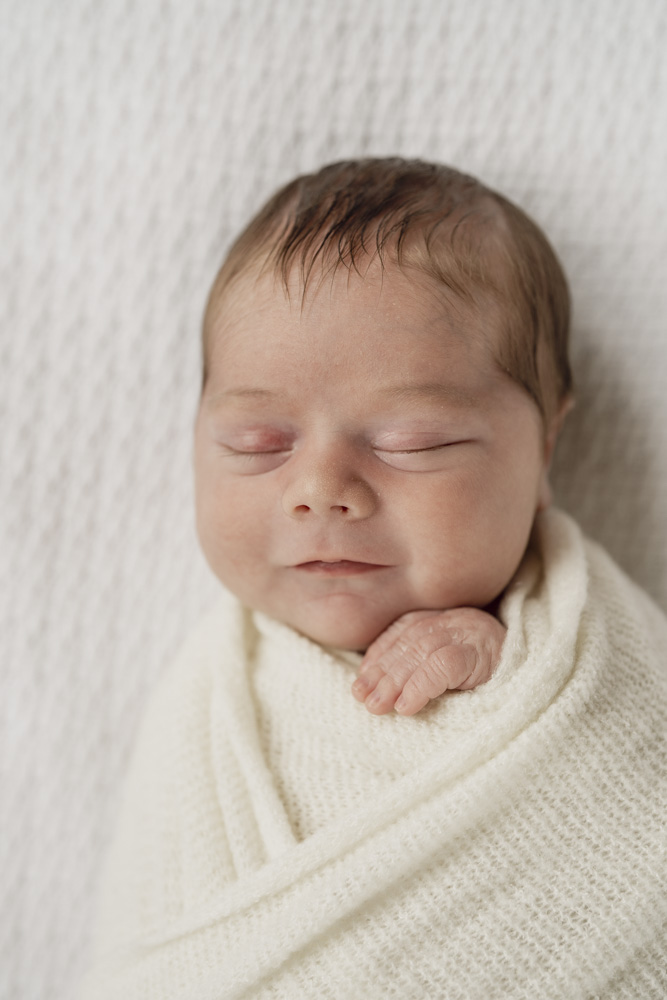 Baby Photography, close up of a smiling newborn swaddled in a beige blanket with one hand sticking out eyes closed