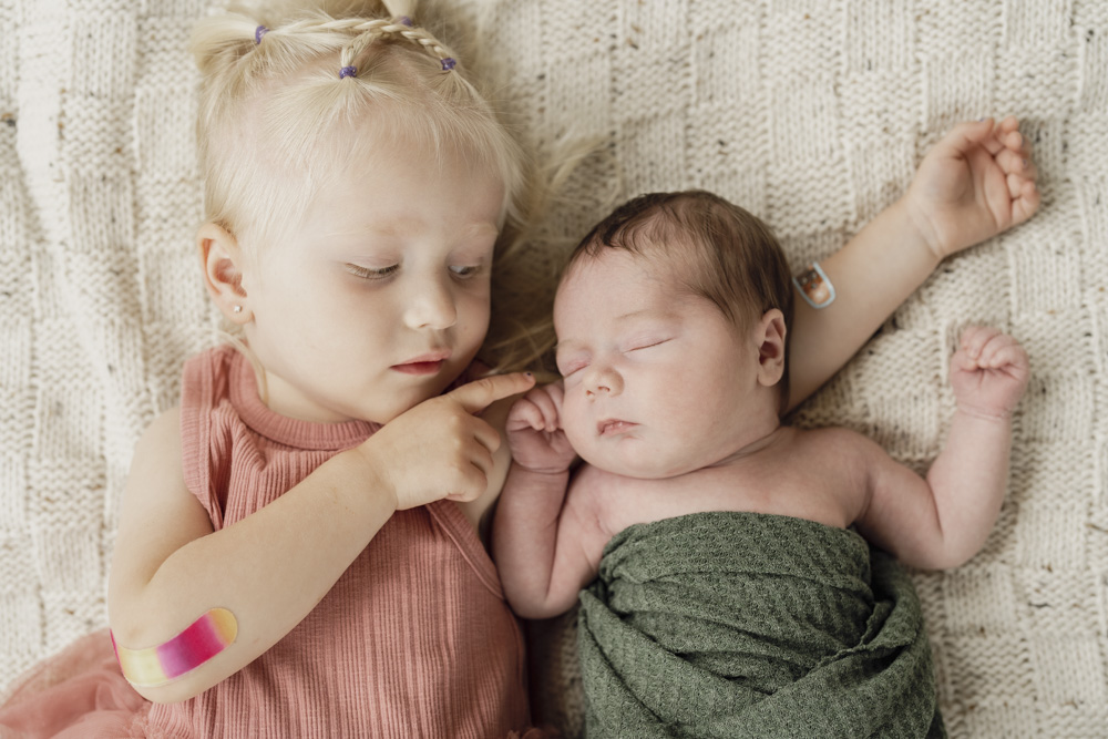 Family Session, sleeping newborn swaddled in a green blanket lying next to big sister on a beige blanket