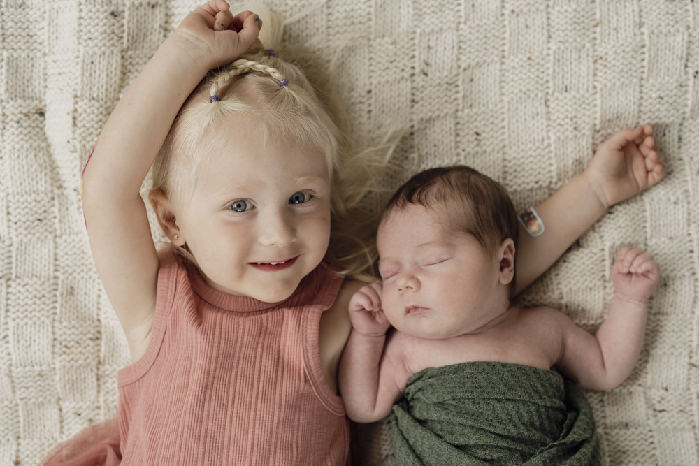 Family Session, sleeping newborn swaddled in a green blanket lying next to big sister on a beige blanket while she is smiling at the camera