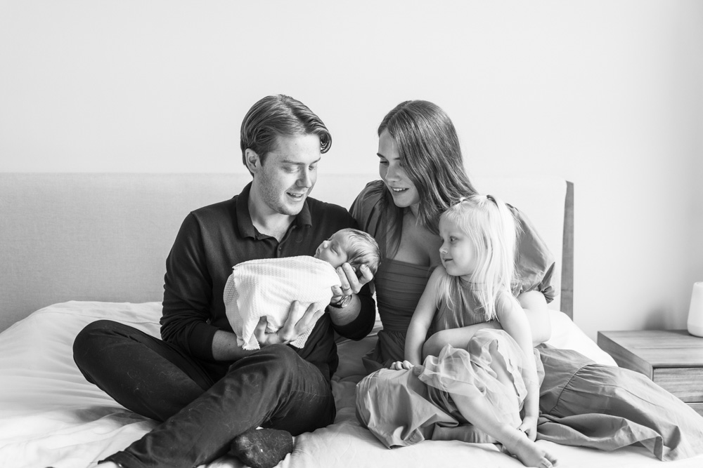 Family Photography, black and white photo of a family of four sitting on the bed while father is holding the newborn baby and the big sister is leaning on mom while all are looking at the baby