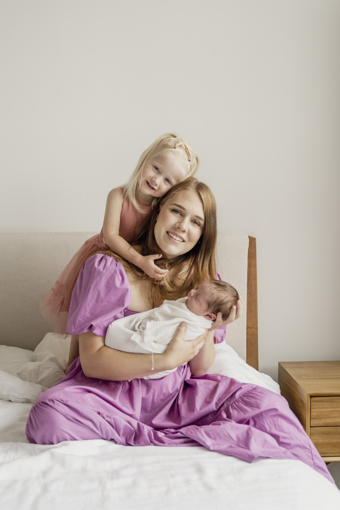 Family Session, mom in a beautiful pink dress is sitting on the bed holding her newborn baby while the big sister is standing behind her mom with her arms wrapped around her
