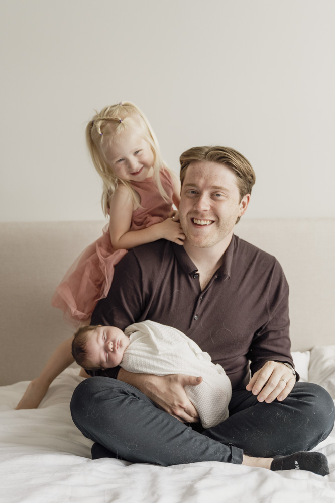 Family Session, father is sitting on the bed holding her newborn baby while the big sister is standing behind him smiling