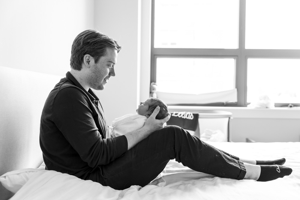 Newborn Photography, black and white photo of father sitting on the bed holding the sleeping newborn baby