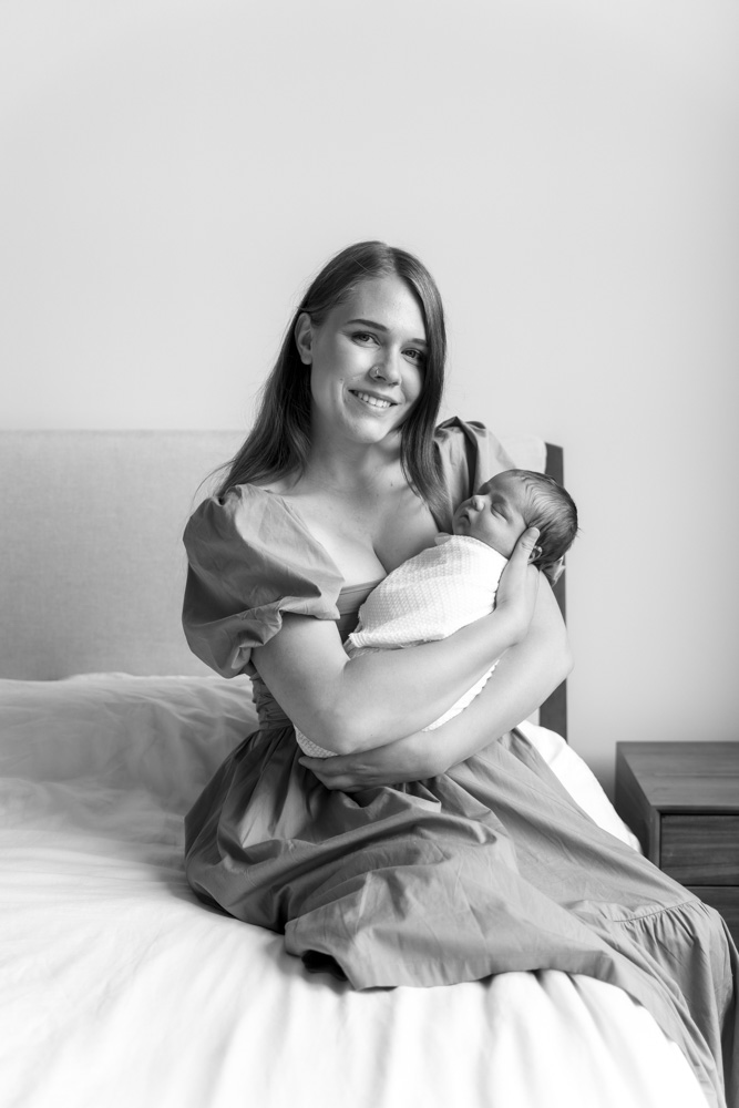 Newborn Photography, black and white photo of mother sitting on the bed holding her sleeping newborn baby smiling at the camera