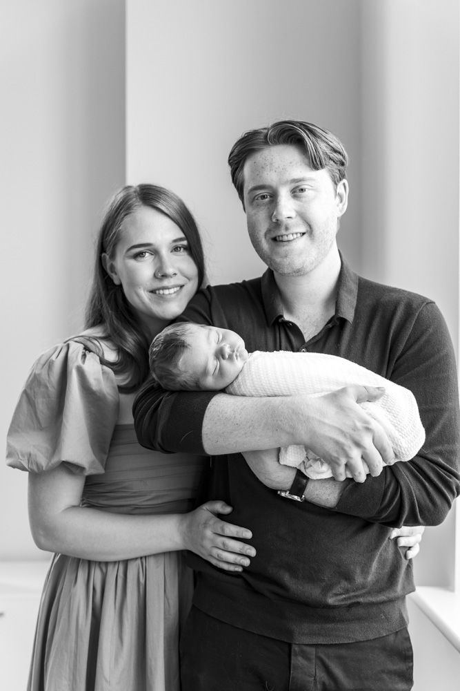 Family Session, black and white photo of parents standing next to a window while father is holding the sleeping newborn baby