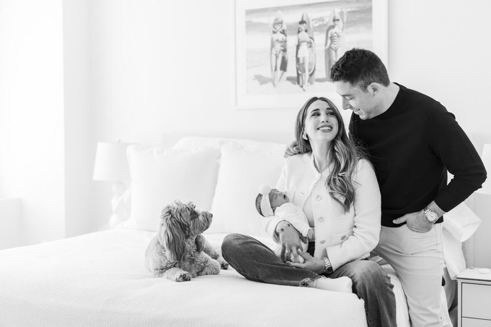 Family Session, black and white photo of mother sitting next do their dog on the bed holding her sleeping newborn baby while father is putting his arm around her and smiling at her, baby is swaddled in a blanket and is wearing a headband with a big bow