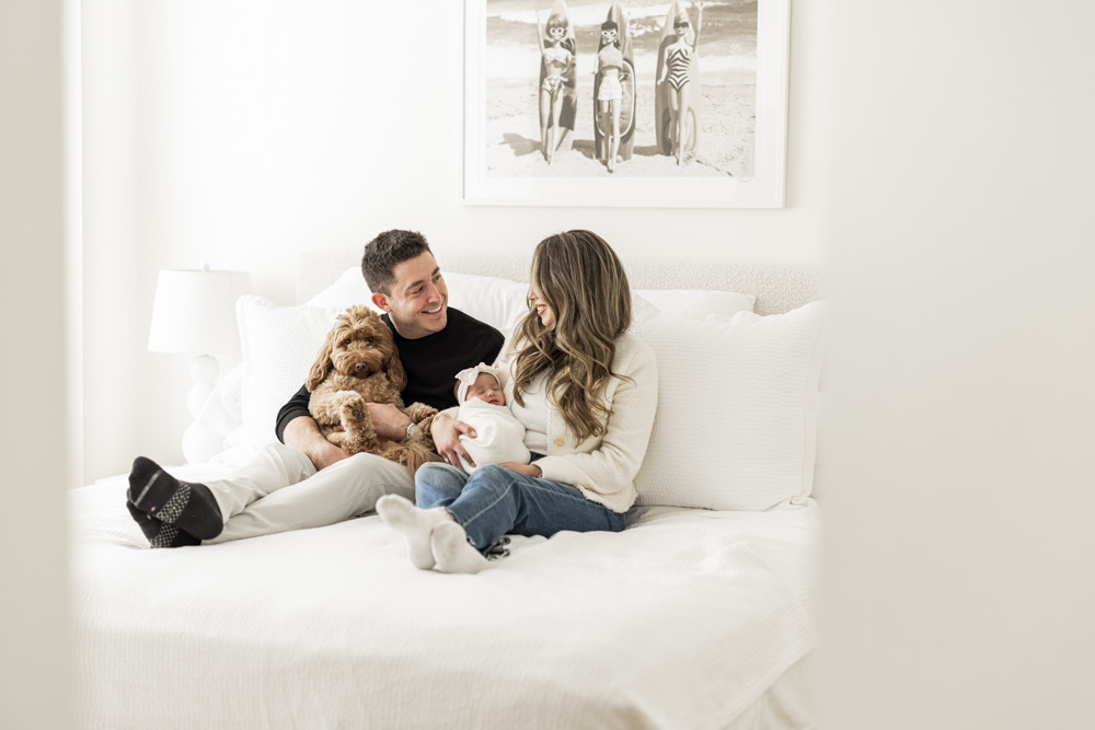 Family Session, parents sitting on the bed smiling at each other while mom is holding their sleeping newborn baby girl and father is holding their cute brown dog