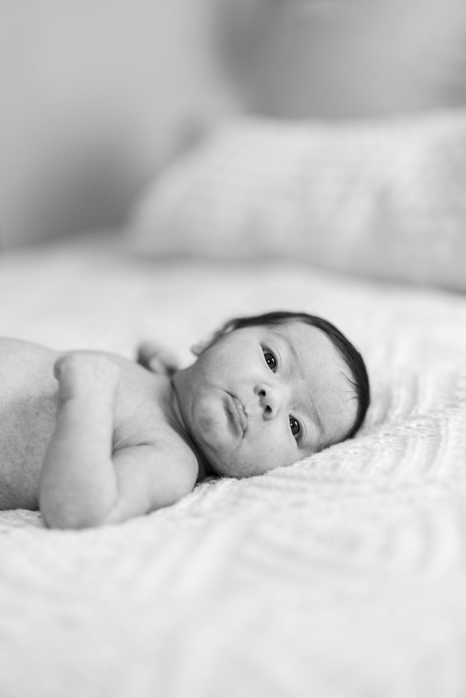 Newborn Photography, black and white photo of newborn baby lying on the bed looking at the camera