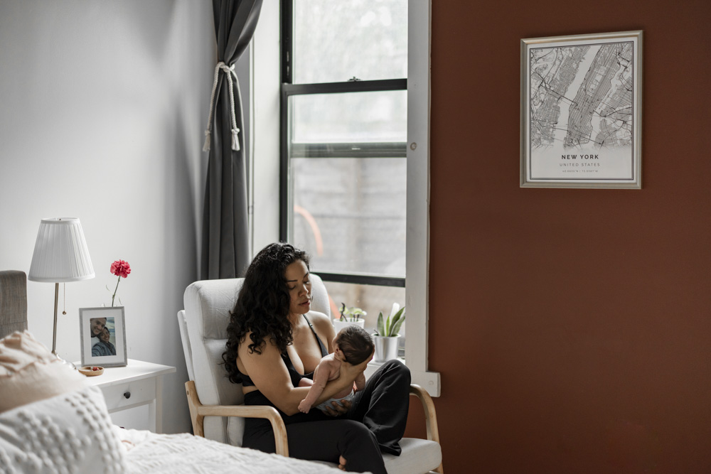 Baby Session, mom is sitting in a chair next to the window holding her newborn baby in front of her