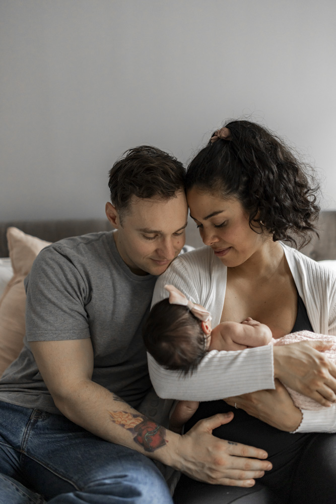 Baby Photography, proud parents sitting next to each other on the couch while mom is holding her newborn baby, both are smiling at the baby, baby has dark hair in is wearing a bow