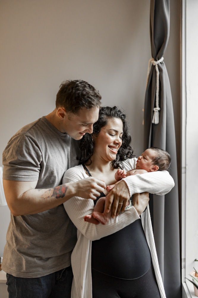 Family Photos, parents are standing next to a window mom is holding her sleeping newborn baby while both are smiling at the baby