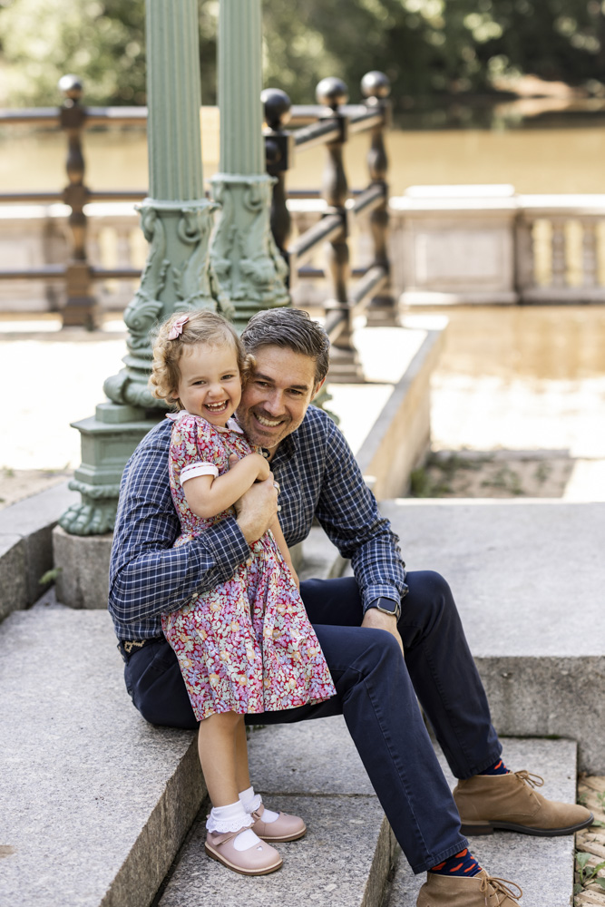 Family Session, father is sitting on steps outside with his arm wrapped around his smiling daughter wearing a beautiful dress both are laughing at the camera